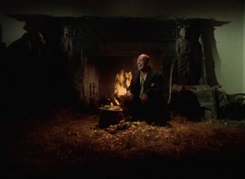 Movie still from “The Tempest” (1979), directed by Derek Jarman – A man sitting in front of a fire in front of a fireplace; Wide shot, High angle