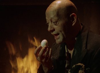 Movie still from “The Tempest” (1979), directed by Derek Jarman – A bald man holding an egg in front of a fireplace; Close Up shot, Low angle