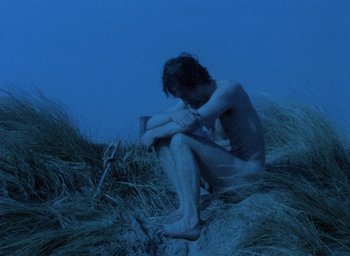 Movie still from “The Tempest” (1979), directed by Derek Jarman – A man sitting on top of a grass covered hill; Wide shot, High angle