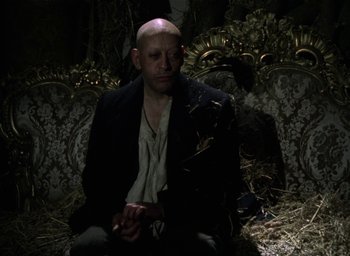 Movie still from “The Tempest” (1979), directed by Derek Jarman – A bald man sitting on a couch in a dark room; Medium shot, High angle