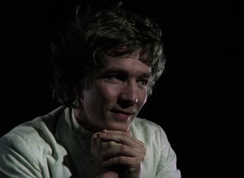 Movie still from “The Tempest” (1979), directed by Derek Jarman – A man with curly brown hair is sitting in the dark with his hands clasped together; Close Up shot, Low angle