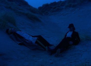 Movie still from “The Tempest” (1979), directed by Derek Jarman – Two people laying in the sand at the beach; Wide shot, High angle