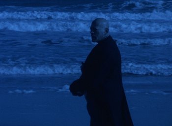 Movie still from “The Tempest” (1979), directed by Derek Jarman – An older man walking on the beach near the ocean; Wide shot, Low angle