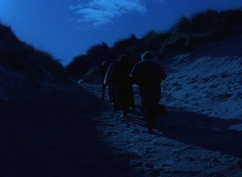 Movie still from “The Tempest” (1979), directed by Derek Jarman – A group of people walking up a hill at night; Extreme Wide shot, High angle