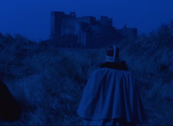 Movie still from “The Tempest” (1979), directed by Derek Jarman – A person wearing a cape standing in a field at night with a castle in the background; Extreme Wide shot, Over the shoulder angle
