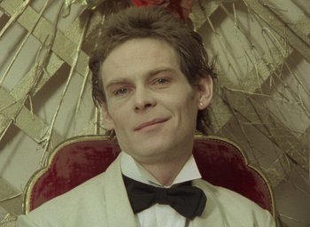 Movie still from “The Tempest” (1979), directed by Derek Jarman – A man in a bow tie sitting in front of an umbrella; Close Up shot, Low angle