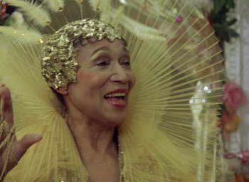 Movie still from “The Tempest” (1979), directed by Derek Jarman – An older woman wearing a yellow dress and a gold hat; Close Up shot, Low angle