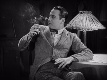 Movie still from “The Ten Commandments” (1923), directed by Cecil B. DeMille – A man sitting in a chair smoking a cigarette and holding a gun; Medium shot, Low angle