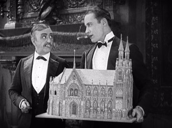 Movie still from “The Ten Commandments” (1923), directed by Cecil B. DeMille – Two men in tuxedos holding a model of a church; Medium shot, Low angle