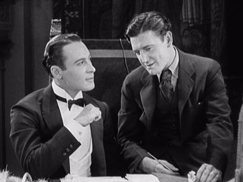 Movie still from “The Ten Commandments” (1923), directed by Cecil B. DeMille – Two men are sitting at a table and one of the men is holding a tie; Medium shot, High angle