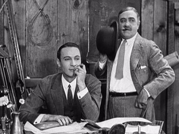 Movie still from “The Ten Commandments” (1923), directed by Cecil B. DeMille – A man sitting at a table with another man standing next to him; Medium shot, Low angle