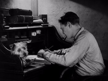 Movie still from “The Ten Commandments” (1923), directed by Cecil B. DeMille – A man sitting at a desk writing on a piece of paper with a dog sitting next to him; Medium shot, High angle