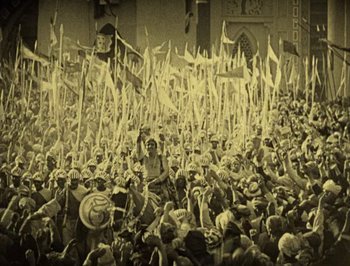 Movie still from “The Thief of Bagdad” (1924), directed by Raoul Walsh – An old photo of a crowd of people holding flags; Extreme Wide shot, High angle