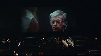 Movie still from “The Thin Blue Line” (1988), directed by Errol Morris – An old man in glasses is on a big screen; Medium shot, Over the shoulder angle