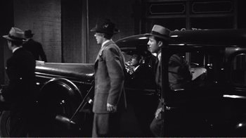 Movie still from “The Thin Blue Line” (1988), directed by Errol Morris – A man and a woman in a suit and hat next to an old car; Wide shot, Over the shoulder angle