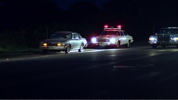 Movie still from “The Thin Blue Line” (1988), directed by Errol Morris – A police car and another car on the side of the road at night; Wide shot, Low angle