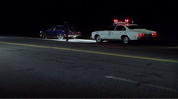 Movie still from “The Thin Blue Line” (1988), directed by Errol Morris – A police car is parked on the side of the road; Wide shot, Low angle