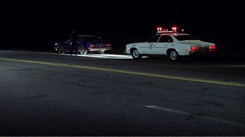 Movie still from “The Thin Blue Line” (1988), directed by Errol Morris – A police officer standing next to a police car on the side of the road; Wide shot, Low angle