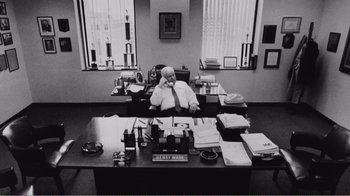 Movie still from “The Thin Blue Line” (1988), directed by Errol Morris – An older man sitting at a desk in an office; Wide shot, High angle