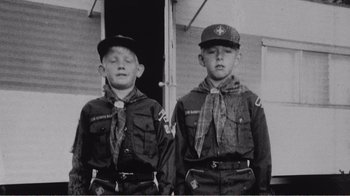 Movie still from “The Thin Blue Line” (1988), directed by Errol Morris – Two young boys in scout uniforms standing next to each other in front of a building; Medium shot, High angle