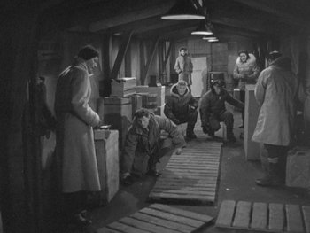 Movie still from “The Thing from Another World” (1951), directed by Howard Hawks – A black and white photo of a group of people in a warehouse; Wide shot, High angle
