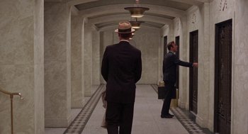 Movie still from “The Thomas Crown Affair” (1968), directed by Norman Jewison – A man in a suit and a hat standing in a hallway with a briefcase; Wide shot, Over the shoulder angle