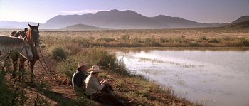 Movie still from “The Three Burials of Melquiades Estrada” (2005), directed by Tommy Lee Jones – A man and a woman sitting next to each other near a body of water; Extreme Wide shot, High angle