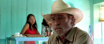 Movie still from “The Three Burials of Melquiades Estrada” (2005), directed by Tommy Lee Jones – An older man wearing a straw hat sitting in front of a bar; Close Up shot, Over the shoulder angle