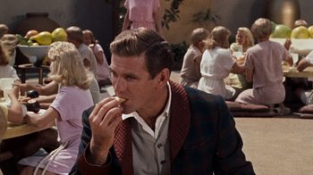 Movie still from “The Time Machine” (1960), directed by George Pal – A man eating something while sitting in front of a group of people; Medium shot, Over the shoulder angle