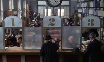 Movie still from “The Tin Drum” (1979), directed by Volker Schlöndorff – An older man wearing a hat is standing in front of a counter; Wide shot, High angle