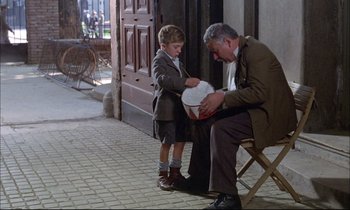 Movie still from “The Tin Drum” (1979), directed by Volker Schlöndorff – An older man sitting on a chair next to a young boy; Wide shot, Over the shoulder angle