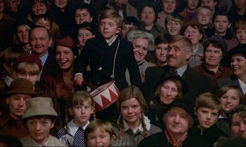 Movie still from “The Tin Drum” (1979), directed by Volker Schlöndorff – A group of people in a crowd with a boy in a drum; Medium shot, High angle