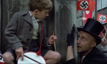 Movie still from “The Tin Drum” (1979), directed by Volker Schlöndorff – A man in a hat and gloves holding a drum and a boy in a suit holding a stick; Medium shot, Over the shoulder angle