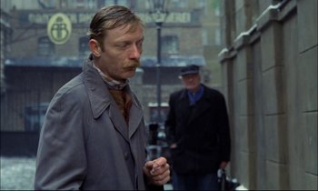 Movie still from “The Tin Drum” (1979), directed by Volker Schlöndorff – A man in a trench coat smoking a cigarette in the rain; Medium shot, Over the shoulder angle