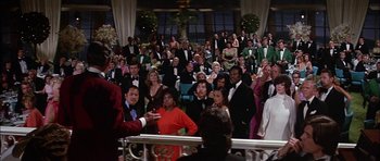 Movie still from “The Towering Inferno” (1974), directed by John Guillermin – A crowd of people in formal wear sitting and standing in a room; Wide shot, Over the shoulder angle
