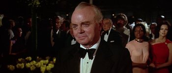 Movie still from “The Towering Inferno” (1974), directed by John Guillermin – An older man wearing a tuxedo in front of a group of people; Close Up shot, Over the shoulder angle