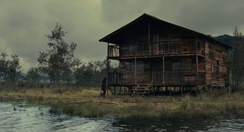 Movie still from “The Towrope” (2012), directed by William Vega – A man standing in front of a house next to a body of water; Extreme Wide shot, Low angle