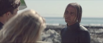 Movie still from “The Tribes of Palos Verdes” (2017), directed by Brendan Malloy – A man standing next to a surfboard on the beach; Close Up shot, Over the shoulder angle