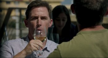 Movie still from “The Trip to Spain” (2017), directed by Michael Winterbottom – A man holding a glass of white wine in front of two other people; Close Up shot, Over the shoulder angle