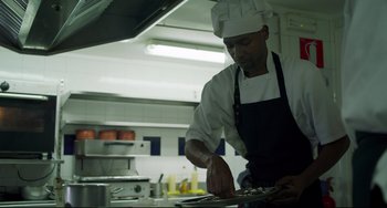 Movie still from “The Trip to Spain” (2017), directed by Michael Winterbottom – A man in a chef's hat and apron preparing food; Medium shot, Low angle