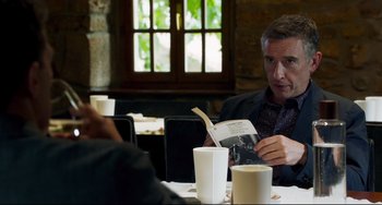 Movie still from “The Trip to Spain” (2017), directed by Michael Winterbottom – A man sitting at a table reading a book; Medium shot, Over the shoulder angle