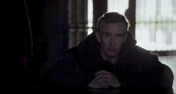 Movie still from “The Trip to Spain” (2017), directed by Michael Winterbottom – A man in a hooded jacket sitting in front of a window; Medium shot, Low angle