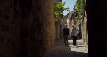 Movie still from “The Trip to Spain” (2017), directed by Michael Winterbottom – A couple of people walking down a street next to a building; Wide shot, Low angle