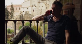 Movie still from “The Trip to Spain” (2017), directed by Michael Winterbottom – A man sitting on a balcony while talking on a cell phone; Medium shot, Low angle