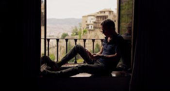 Movie still from “The Trip to Spain” (2017), directed by Michael Winterbottom – A man sitting on a window ledge looking at his cell phone; Wide shot, Low angle