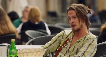Movie still from “The Trip to Spain” (2017), directed by Michael Winterbottom – A man sitting at a table holding a guitar; Medium shot, Over the shoulder angle