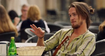 Movie still from “The Trip to Spain” (2017), directed by Michael Winterbottom – A man sitting at a table with a guitar in his hand; Medium shot, Over the shoulder angle