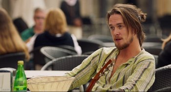 Movie still from “The Trip to Spain” (2017), directed by Michael Winterbottom – A man sitting at a table holding a guitar; Medium shot, Over the shoulder angle
