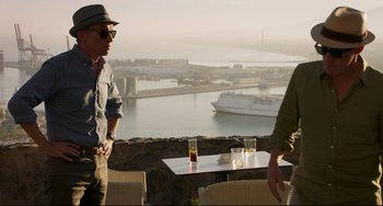 Movie still from “The Trip to Spain” (2017), directed by Michael Winterbottom – Two men are standing at a table overlooking the water; Medium shot, Over the shoulder angle