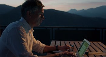 Movie still from “The Trip to Spain” (2017), directed by Michael Winterbottom – A man sitting at a wooden table with a laptop; Medium shot, High angle